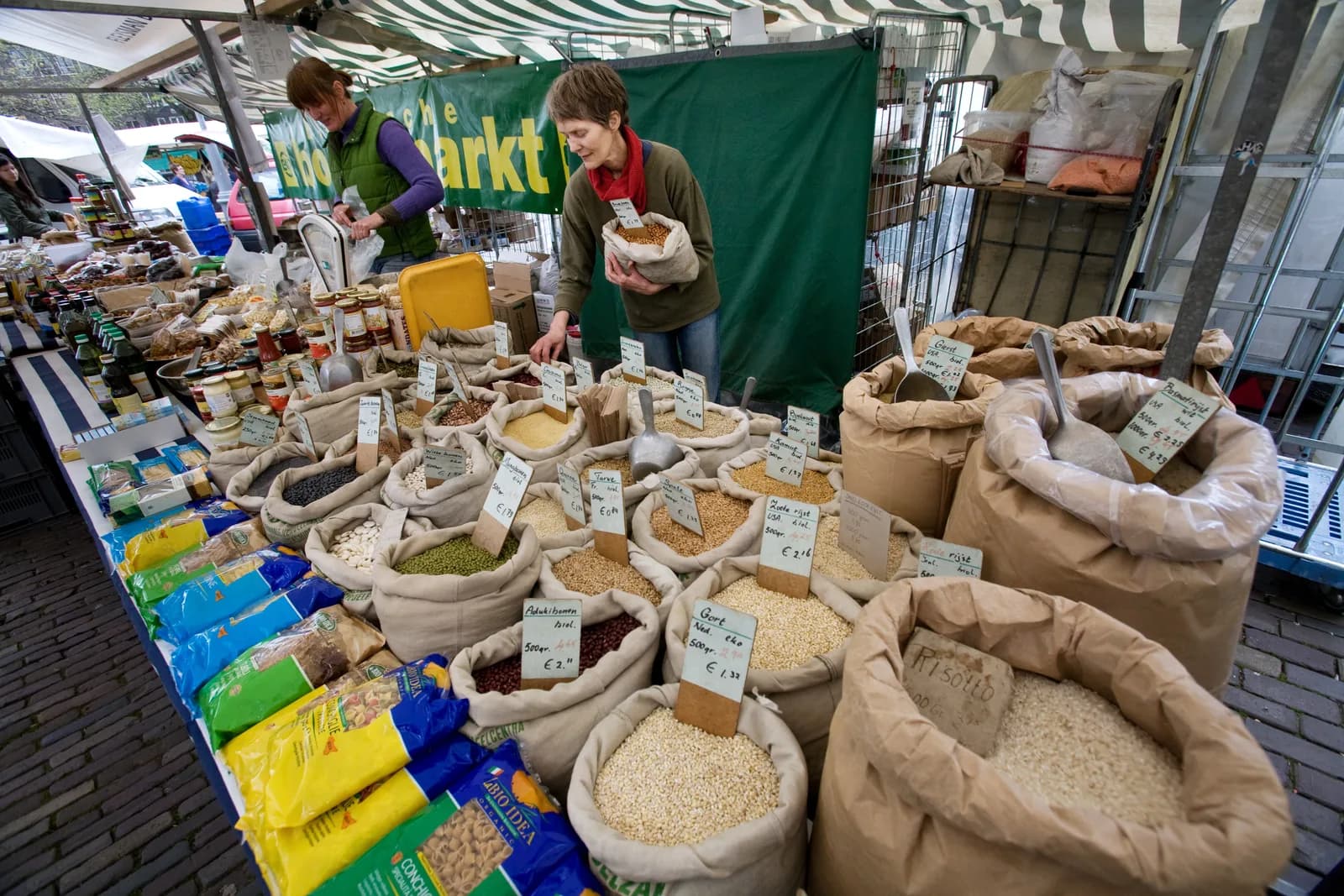 Marktkooplui in Amsterdam wegen producten af tussen zakken granen en peulvruchten, onder een gestreepte luifel.